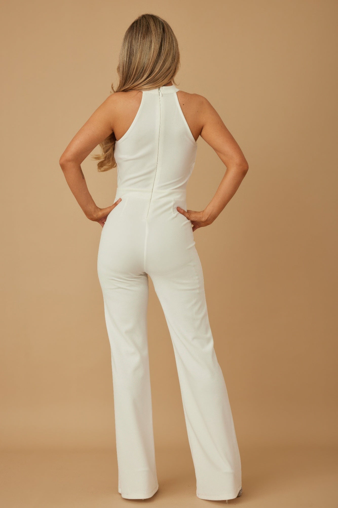 Back view of model wearing white halter chain jumpsuit with zip back fitted bodice and wide leg trousers