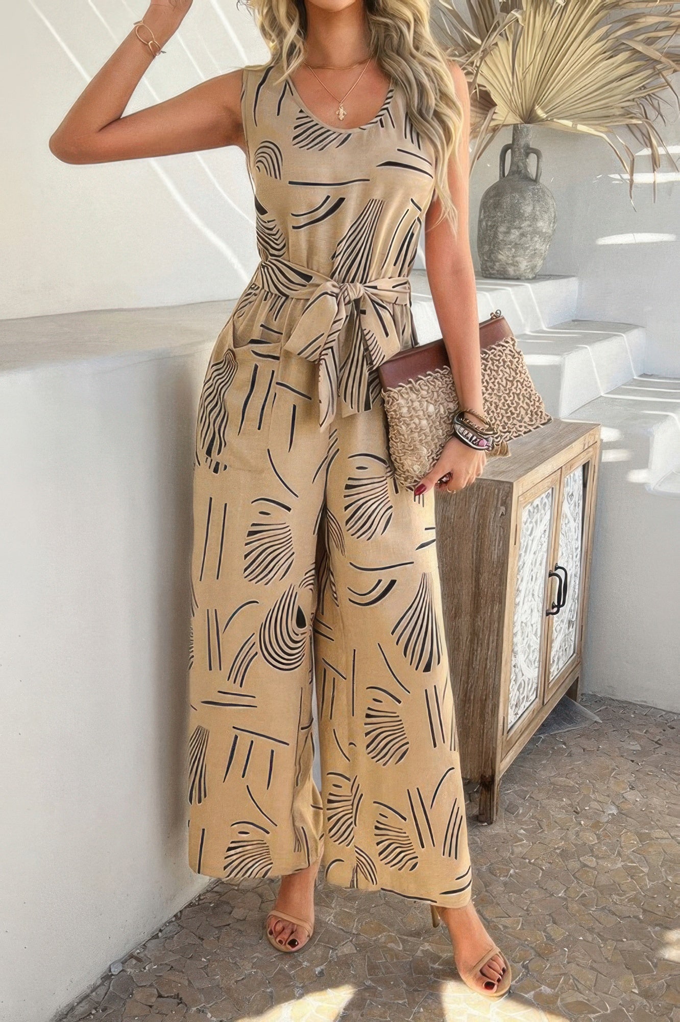 Tie Waist Jumpsuit beige abstract print sleeveless wide leg with self tie belt and model holding woven clutch