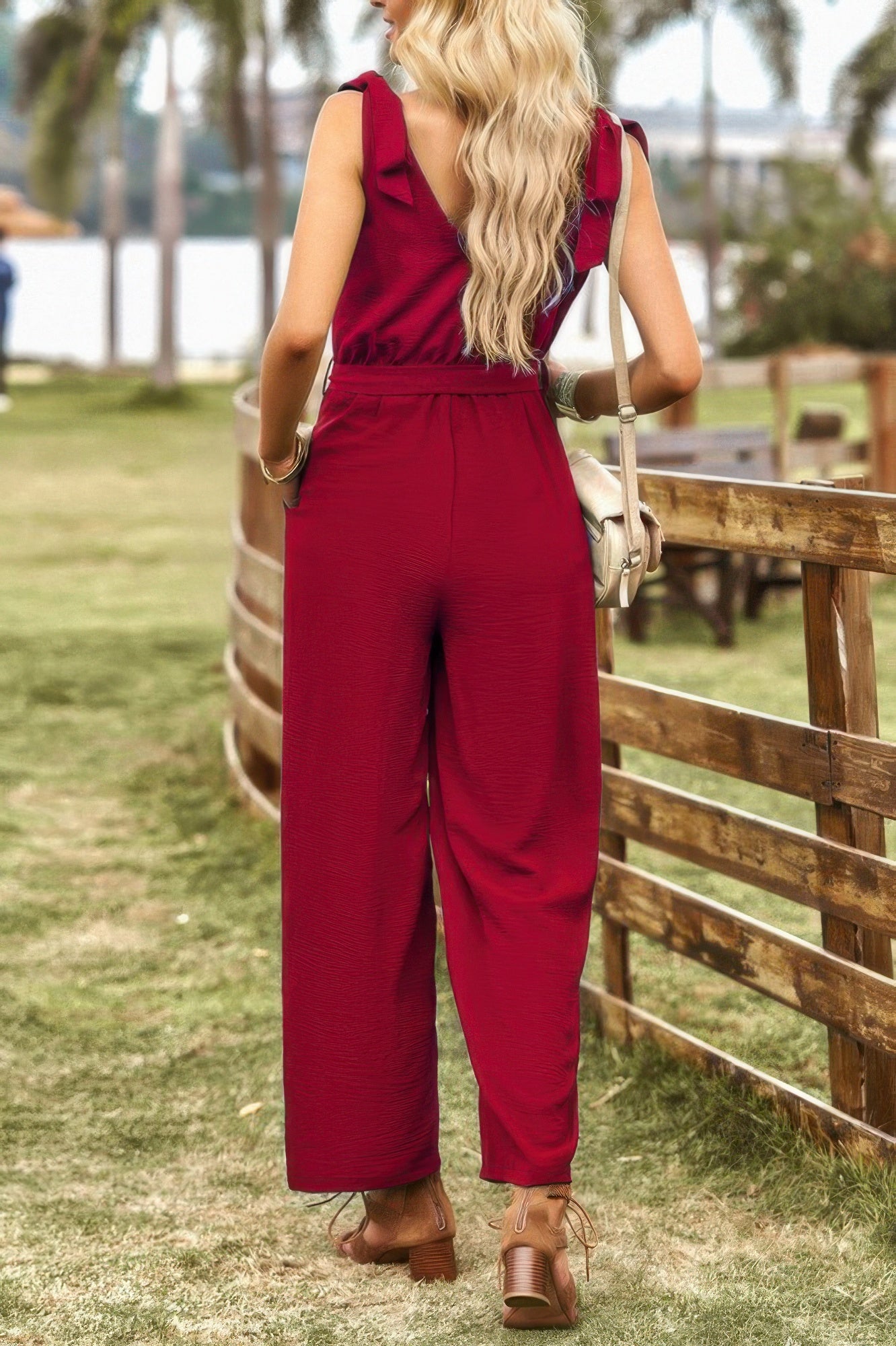 Back view of model wearing CCWHOLESALECLOTHING burgundy women's v neck knot shoulder jumpsuit with elastic waist and sandals