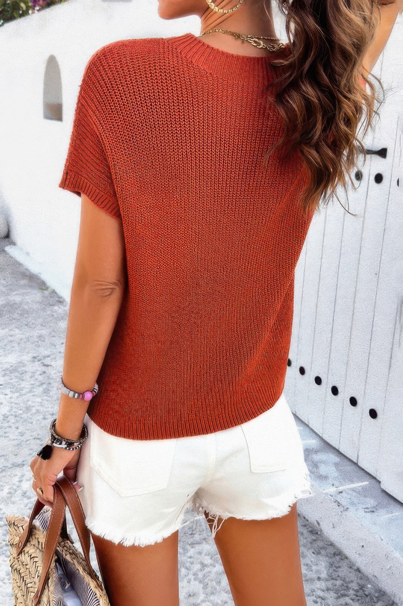 Back view of woman wearing rust ribbed knit loose top with white frayed denim shorts holding straw tote bag