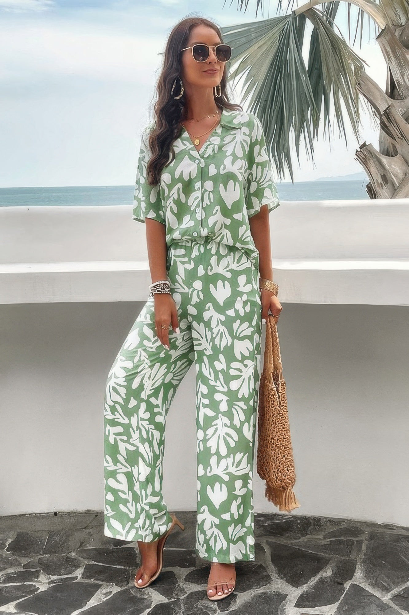 Woman wearing green floral print front button blouse set with wide leg pants straw tote and sunglasses at beach
