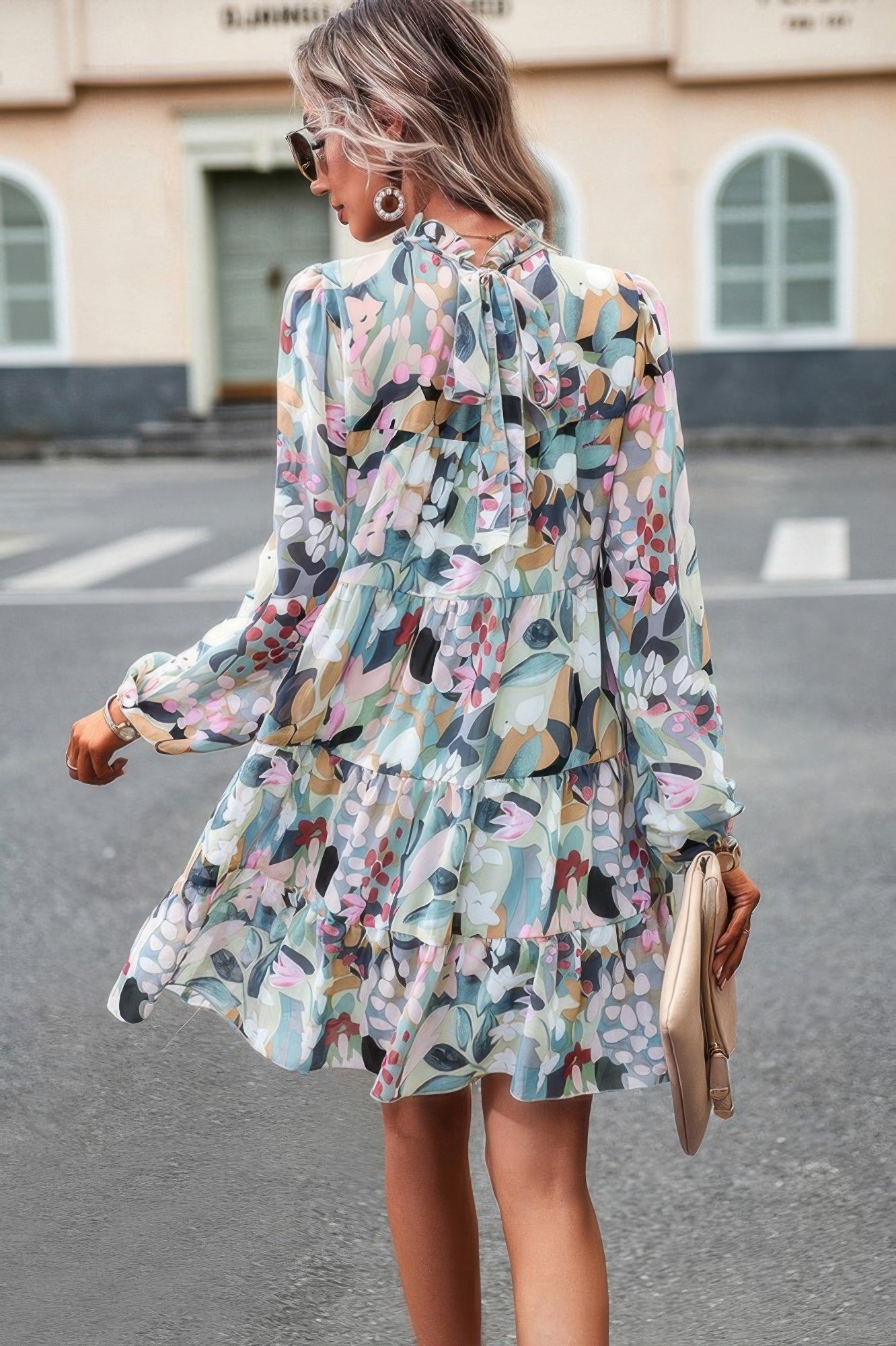 Back view woman wearing CCWHOLESALECLOTHING abstract print tiered dress with long sleeves and neck bow holding beige clutch