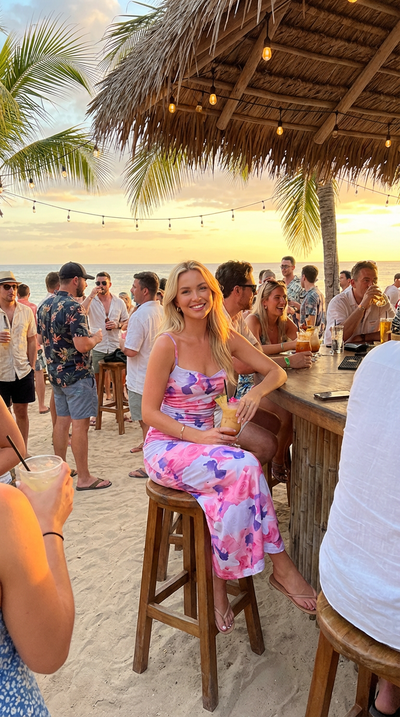 Woman in a colorful dress sitting at a beach bar with sunset and palm trees in the background.
