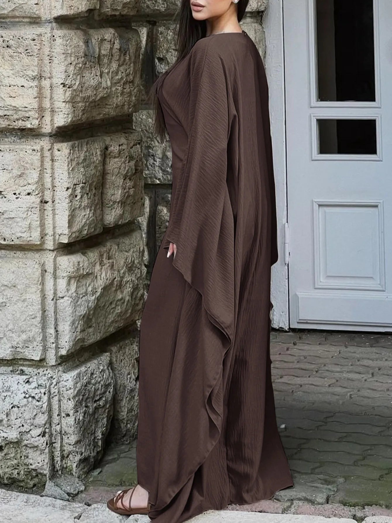Trendsi brown bohemian maxi dress with cape sleeves floor length draped silhouette on model by stone wall
