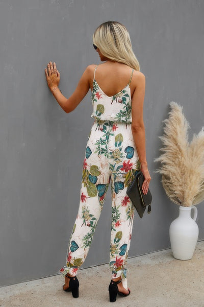 Back view of woman wearing a floral print cami jumpsuit with thin straps holding a clutch purse