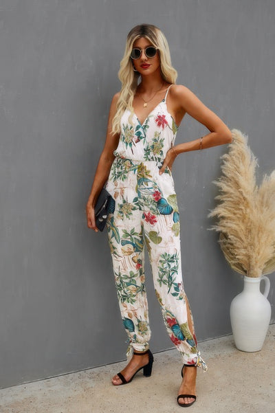 Model wearing white floral print cami jumpsuit with V neckline belted waist tie ankle cuffs and black heels
