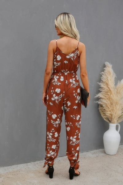 Back view of woman in brown floral print cami jumpsuit with tie ankle black heels and clutch