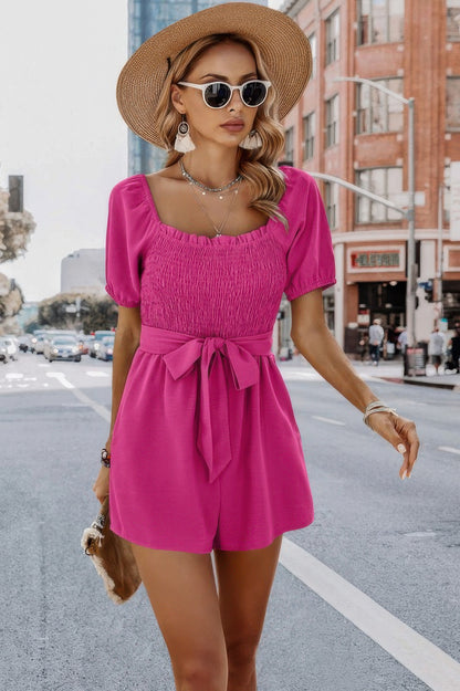 Woman wearing hot pink smocked square neck romper with puff sleeves and tied waist bow straw hat and sunglasses on a city street