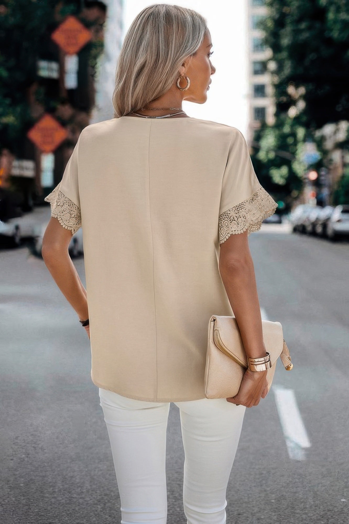Woman wearing beige V Neck Fit Blouse with lace sleeves holding tan clutch and white pants on city street