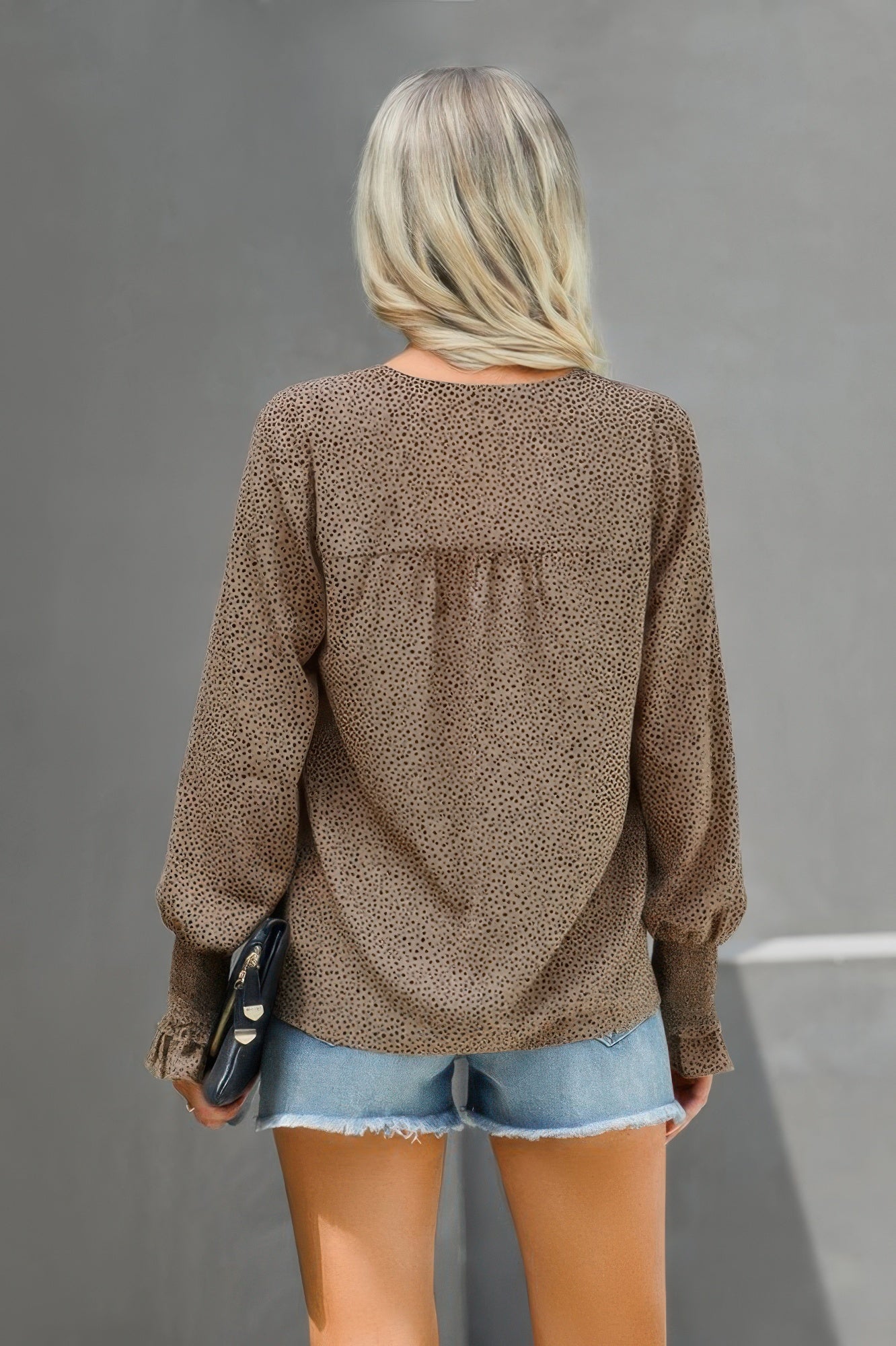 Back view of woman wearing v neck dalmatian print blouse in brown with long smocked sleeves and denim shorts