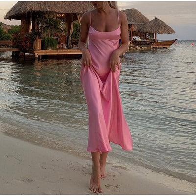 Woman wearing a pink satin slip dress walking barefoot on the beach near water and huts