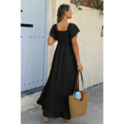 Woman wearing black square neck ruffle hem dress with smocked bodice and tiered skirt carrying straw tote