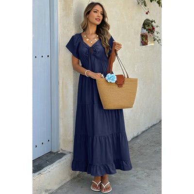 Model wearing navy square neck ruffle hem dress with tiered skirt holding straw tote and sandals
