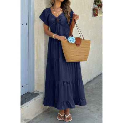 Woman in navy square neck ruffle hem dress with tiered skirt and tie front carrying straw tote and sandals