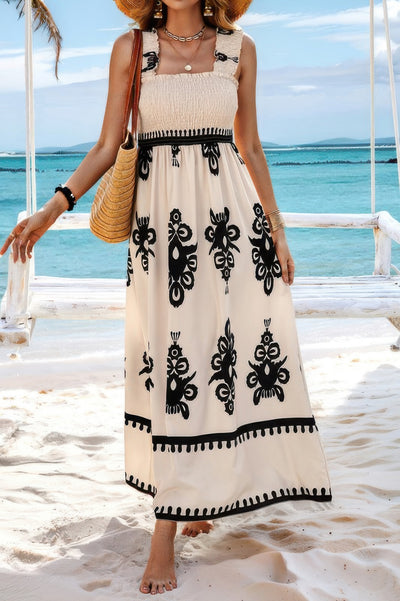 Woman on beach wearing sleeveless square neck dress cream maxi with black floral print smocked bodice and straw bag