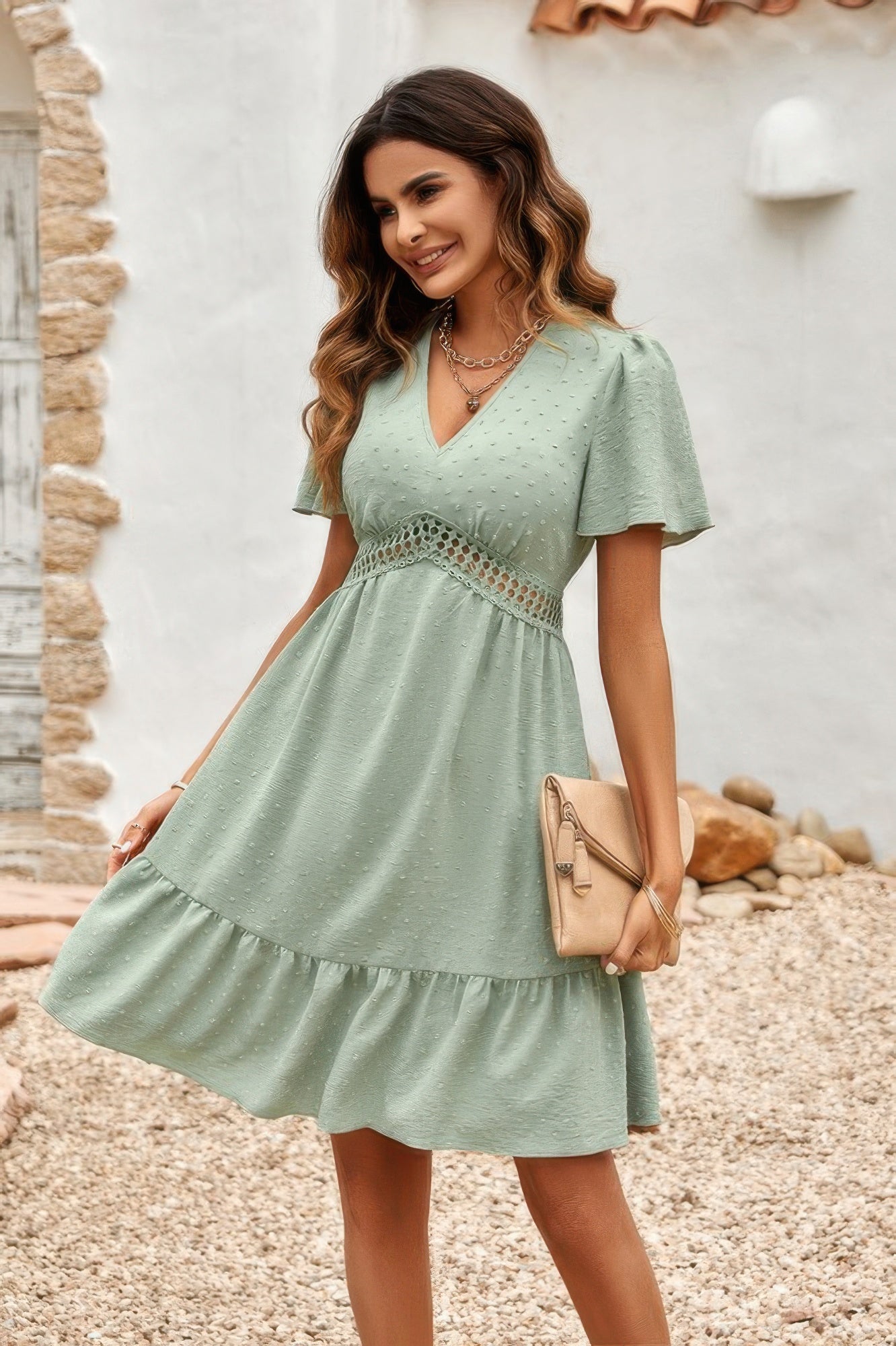 Woman in sage green Swiss dot ruffle dress with V neck flutter sleeves lace waist holding beige clutch outdoors